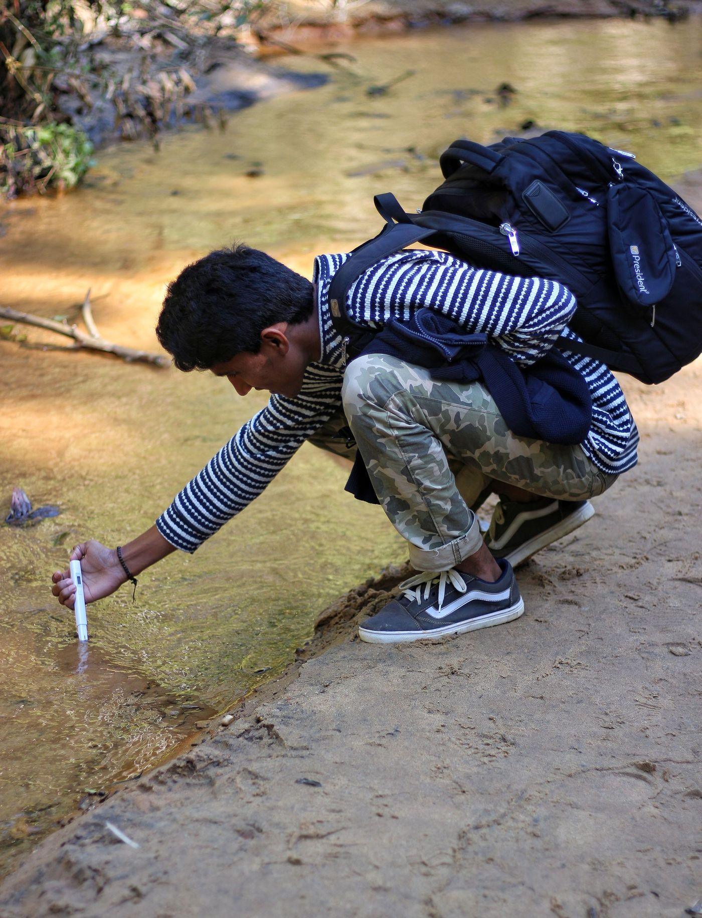 A researcher crouching at the edge of a stream to draw a water sample for field testing